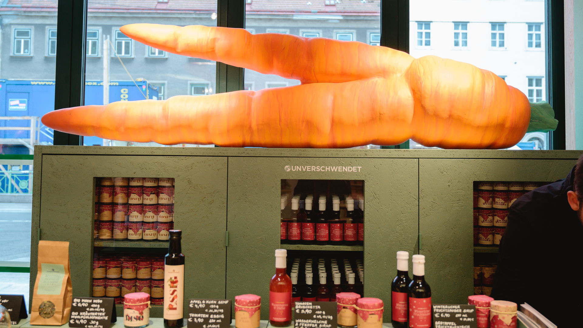 Unverschwendet Ladentresen mit Verkaufsprodukten und riesiger Kunstkarotte im Hintergrund (Vorderansicht) Unverschwendet Ladentresen mit Verkaufsprodukten und riesiger Kunstkarotte im Hintergrund